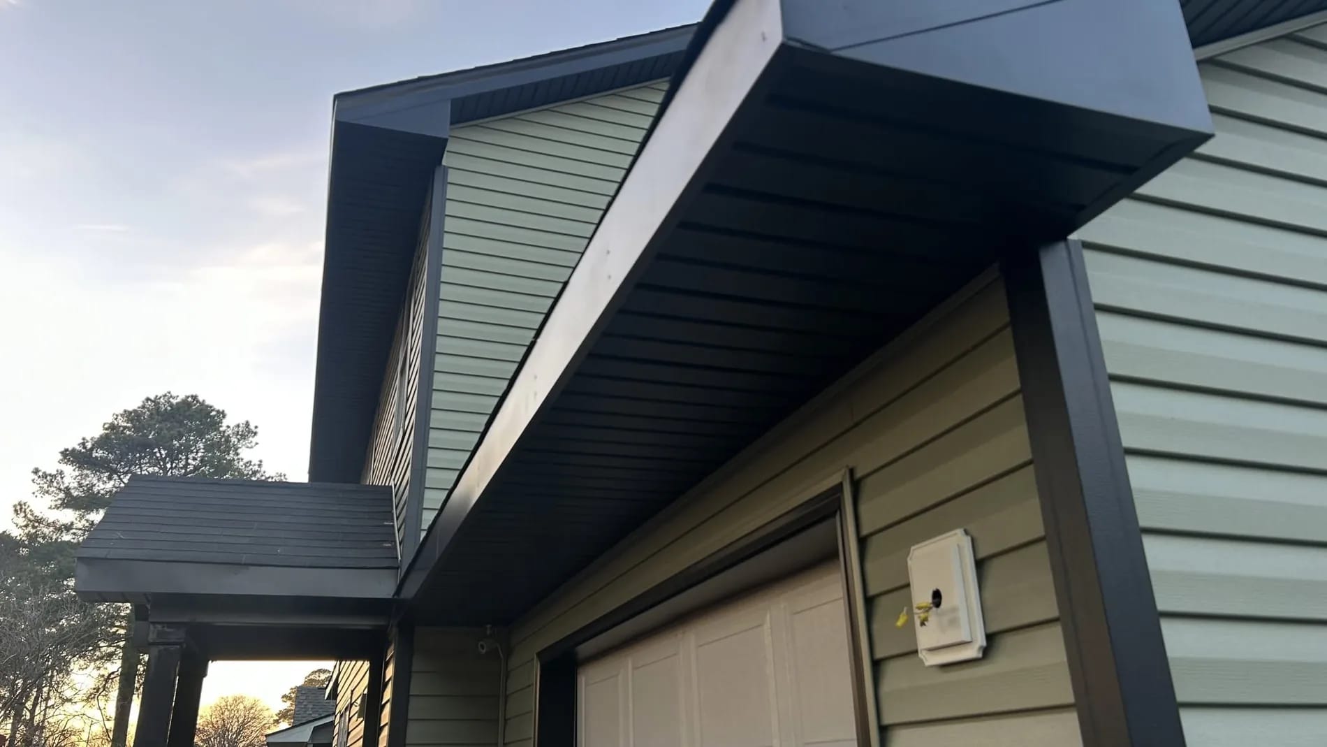 Black soffit and fascia wrap above a garage, over sage-green lap siding, at sunset.